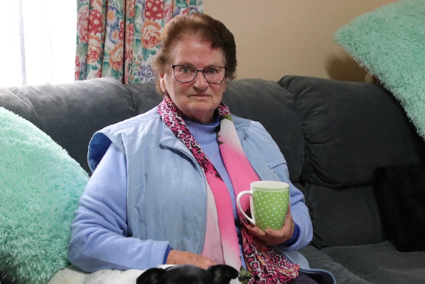 A woman in blue, with a pink scarf, holds a mug as she sits on a grey couch