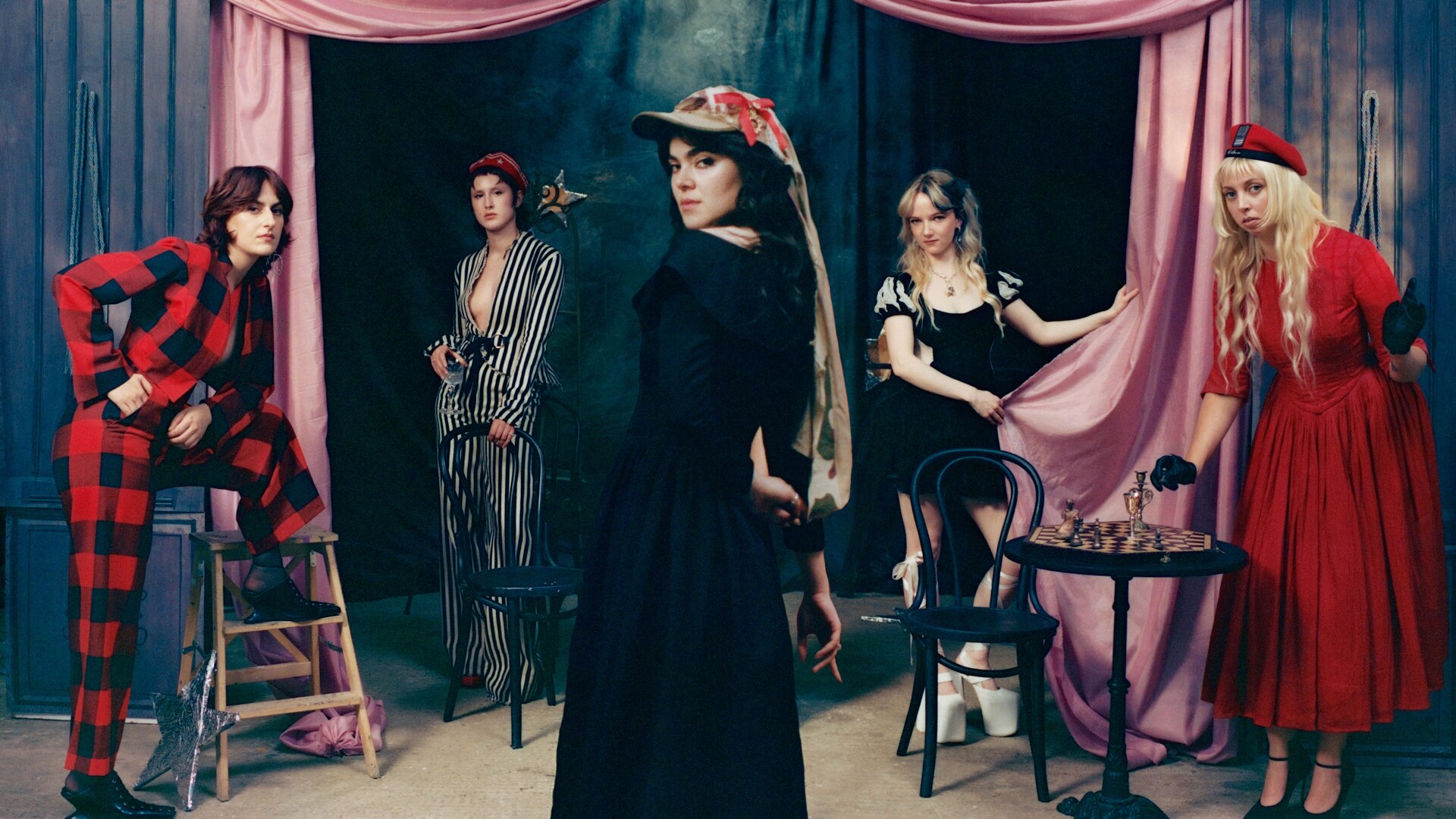 Five femme presenting people stand in a space framed by pink and blue curtains