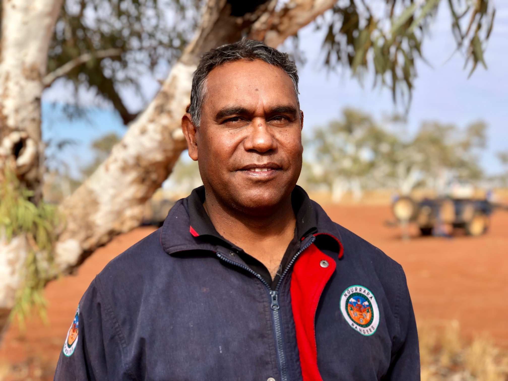 Aboriginal man, Peter Murray, smiling at the camera, he is standing under a tree surrounded by red dessert sand.