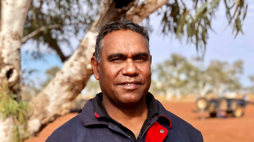 Aboriginal man, Peter Murray, smiling at the camera, he is standing under a tree surrounded by red dessert sand.