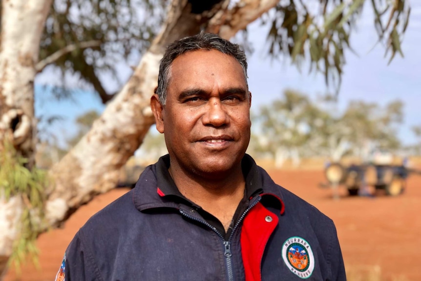 Aboriginal man, Peter Murray, smiling at the camera, he is standing under a tree surrounded by red dessert sand.