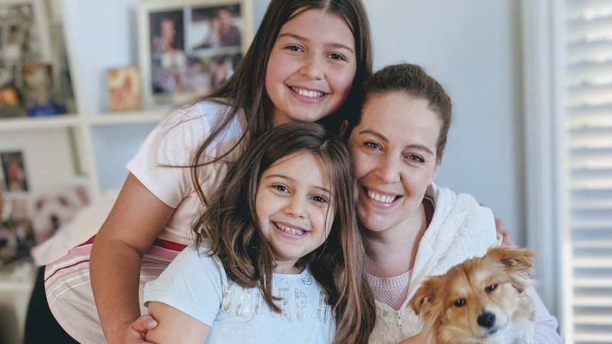 A woman and her young daughters smile for a family photo, while holding a small dog.