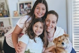 A woman and her young daughters smile for a family photo, while holding a small dog.