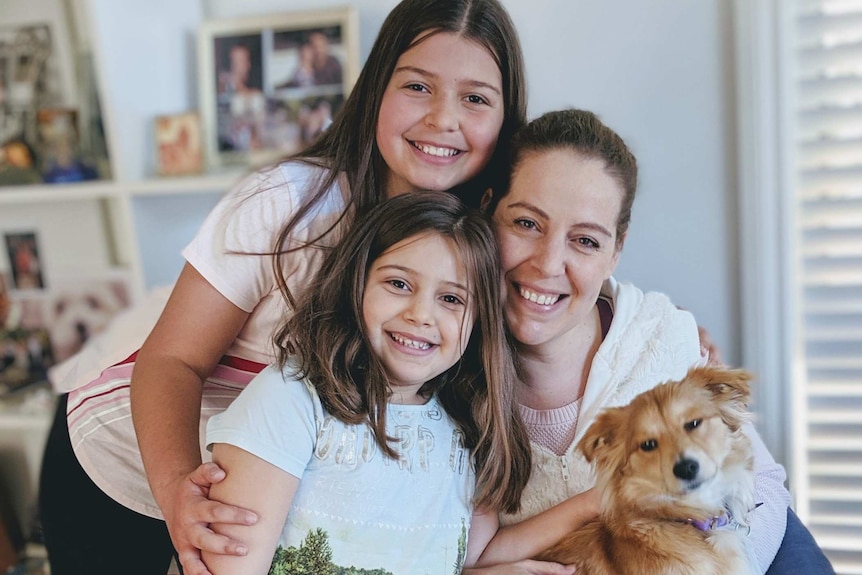 A woman and her young daughters smile for a family photo, while holding a small dog.
