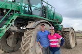 A man and a woman smiling and standing in front of a large green sprayer. 