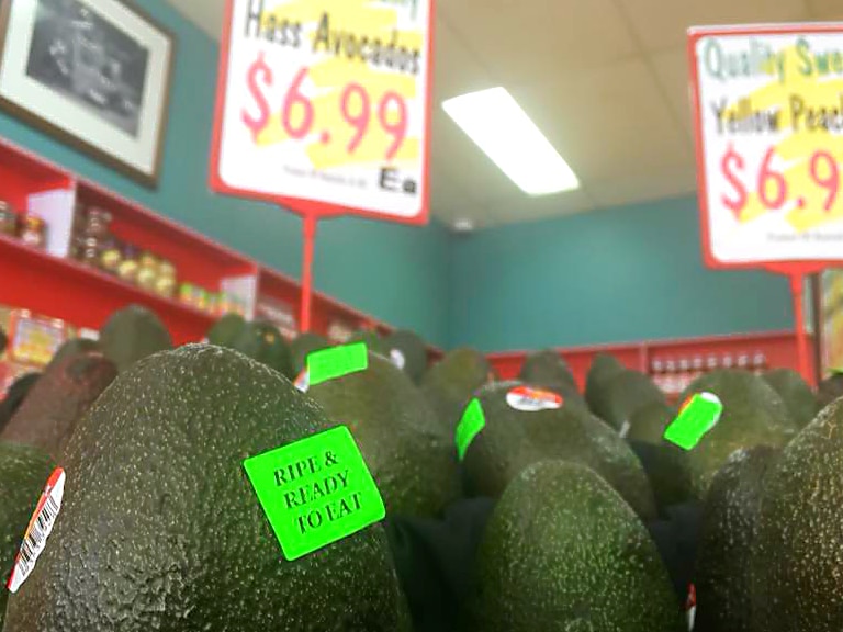 Avocados on a supermarket shelf with the price in the background showing $6.99 each.