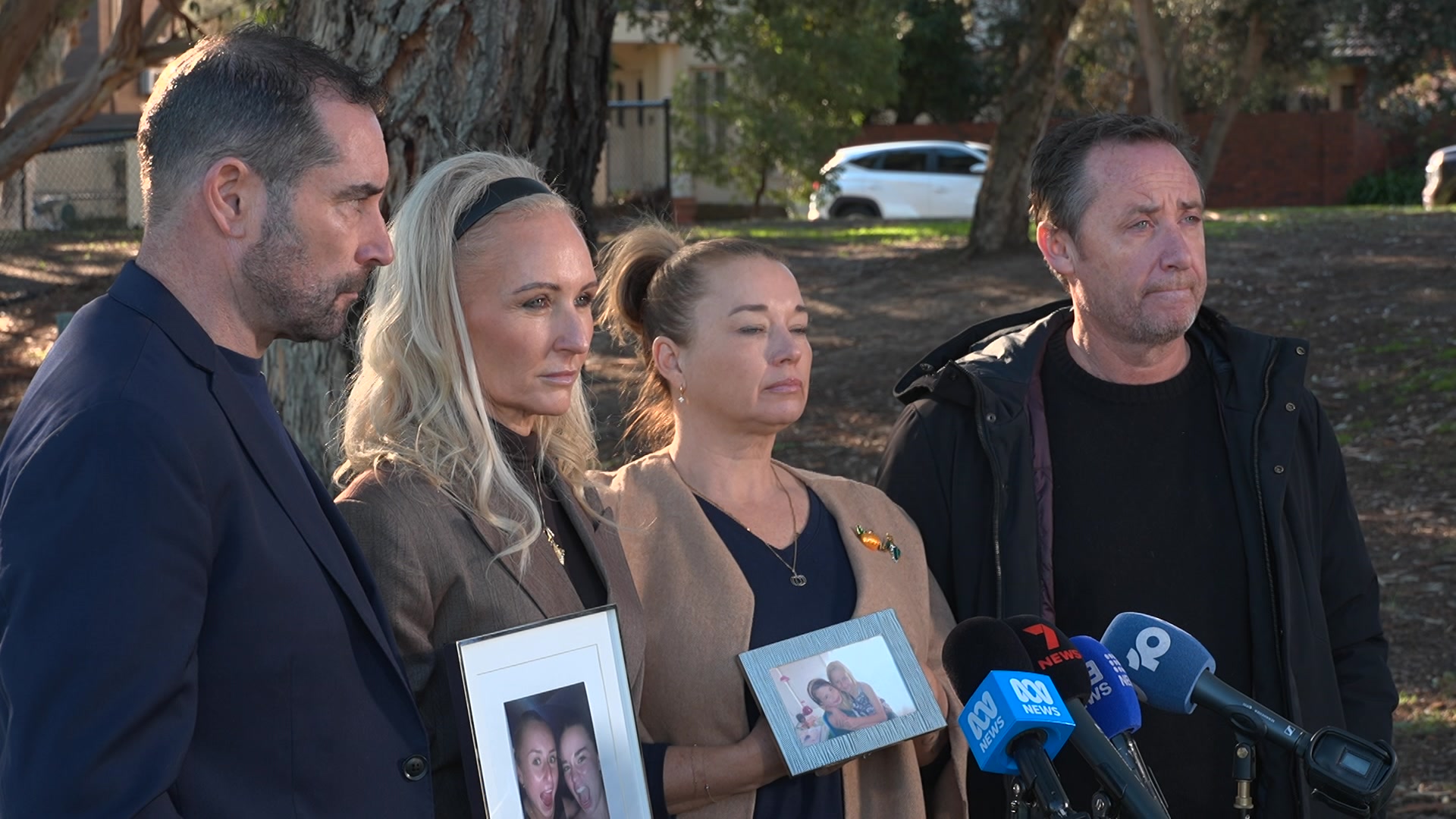 Two  men in dark jackets and shirts booked two women in beige coats and black tops who stand behind microphones.
