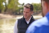 A man wearing a shirt and vest standing in front of a river with a blurred shoulder of another man in the foreground.