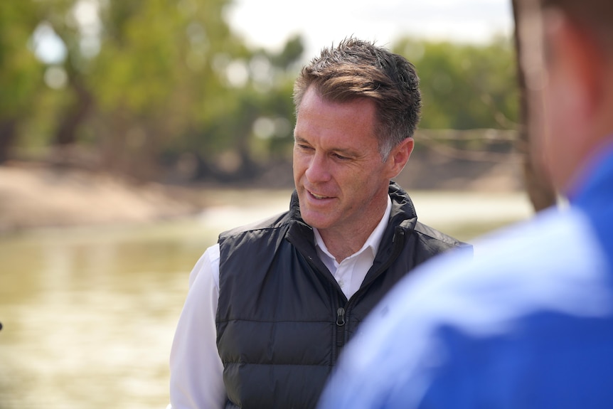 A man wearing a shirt and vest standing in front of a river with a blurred shoulder of another man in the foreground.
