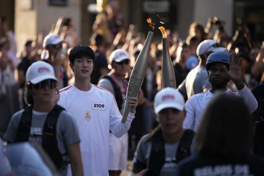 Two men dressed in white carry a large torch through a crowd of people
