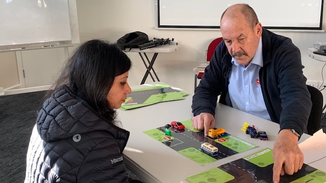 An older man demonstrates road rules to a migrant woman using toy cars.