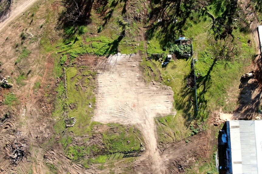 An aerial shot shows a dirt square surrounded by grass where a house has been cleared.
