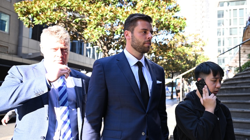 A man in a suit stares ahead outside a court house with another man in a suit next to him on his right.