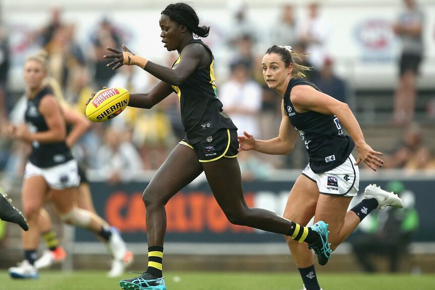 A woman dressed in an AFL uniform and football boots runs with an AFL ball while being chased by another woman.