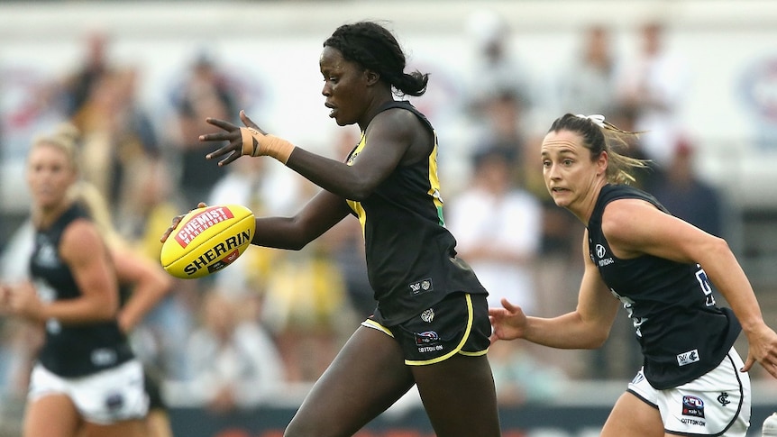 A woman dressed in an AFL uniform and football boots runs with an AFL ball while being chased by another woman.