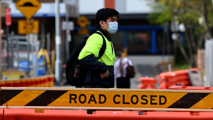 A man wearing a high-vis jumper wears a mask and stands behind a road closed sign.