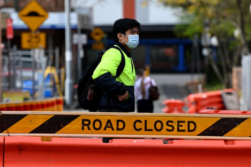 A man wearing a high-vis jumper wears a mask and stands behind a road closed sign.