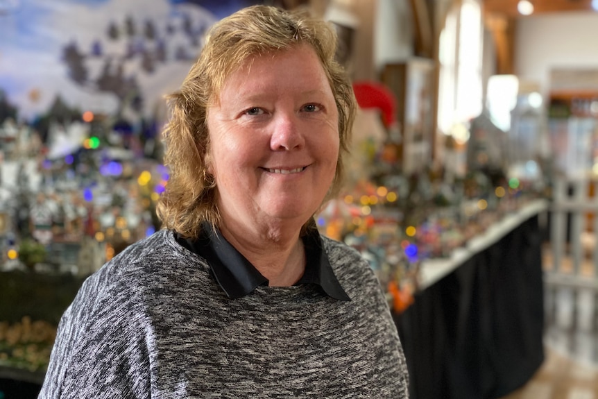 Woman smiles with blurred Christmas lights and decorated village in the background.