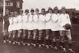 A black and white picture of a women's football team wearing white shirts and black and white hats in front of a goal