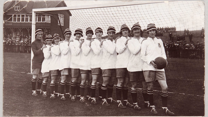A black and white picture of a women's football team wearing white shirts and black and white hats in front of a goal