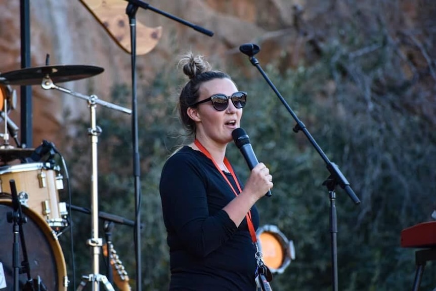 A woman with her hair in a bun, wearing sunglasses and a lanyard, stands on a stage and speaks into a microphone.