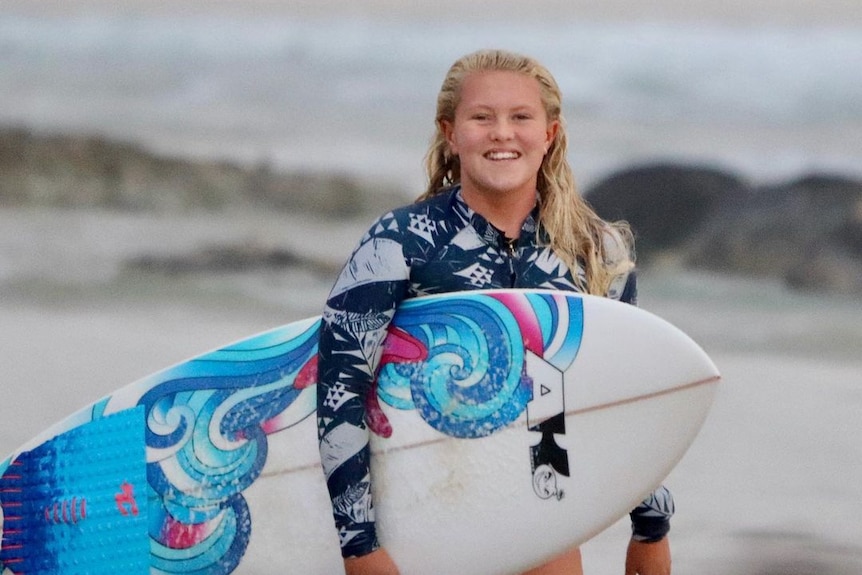 A girl with blonde hair holding a blue and pink and white surfboard