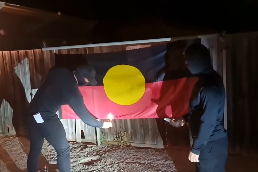 An aboriginal flag hung up with two men wearing all black standing in front of it