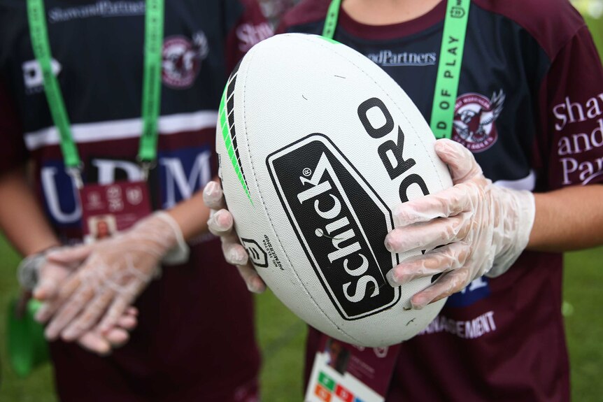 A ball boy holds an NRL rugby league ball while wearing a pair of gloves.