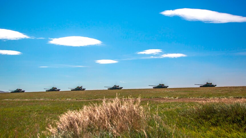 Six tanks move in a line across a green grassy landscape with blue sky and a few small clouds in the background