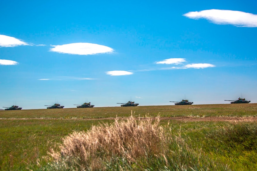 Six tanks move in a line across a green grassy landscape with blue sky and a few small clouds in the background