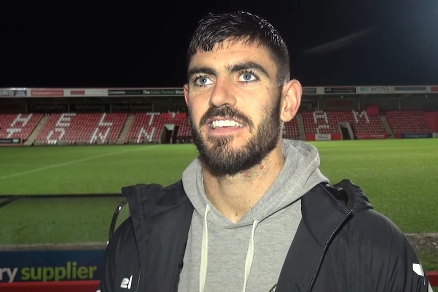 A man wearing a grey hoodie and black coat smiles, standing at the side of a football stadium