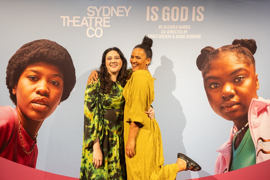 Shari Sebbens and Zindzi Okenyo dressed up in evening wear at a premiere in front of a photo wall with images of Is God Is stars