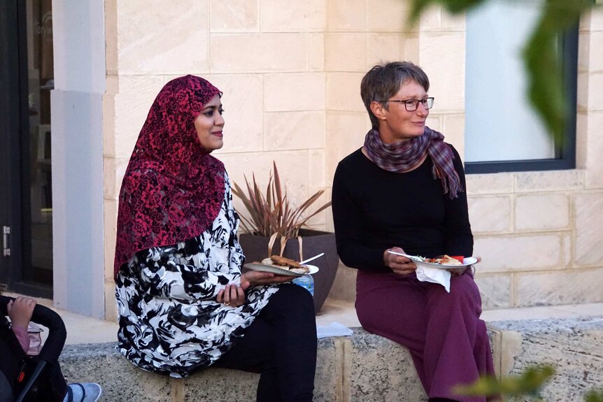 Two women holding plates of food, one wearing a hijab, sit side by side on a brick wall.