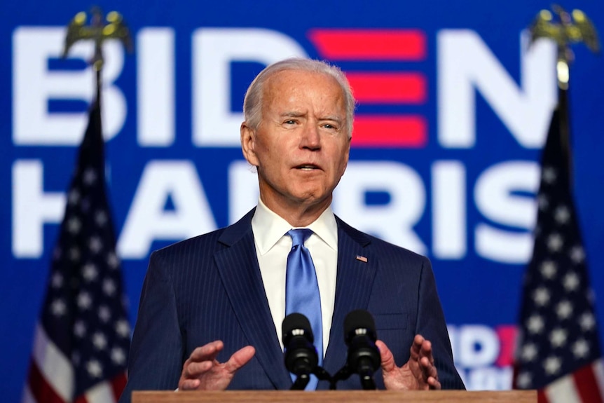 Joe Biden gestures with both hands while speaking at a lectern. He is between two flags and a sign that reads BIDEN HARRIS