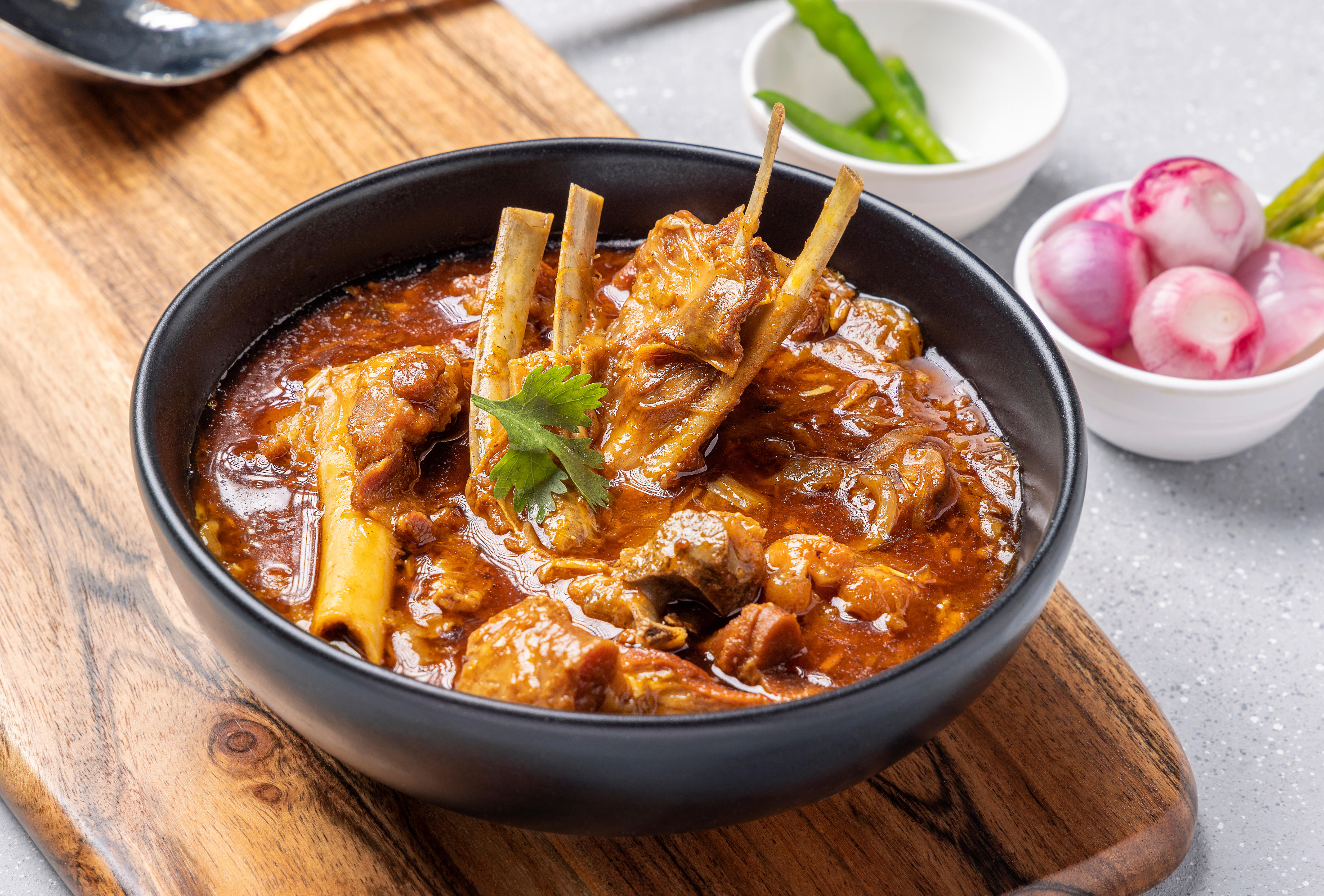 A black bowl of lamb curry in gravy sits on top of a wooden chopping board on a bench.