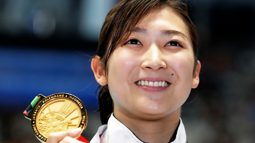 A smiling woman holds up her gold medal.