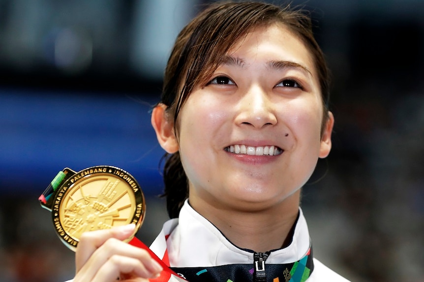 A smiling woman holds up her gold medal.