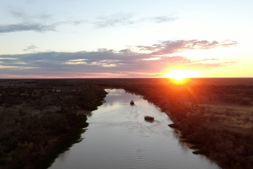 A bright orange sun sets over a long reach of a river with two boats cruising along it. 