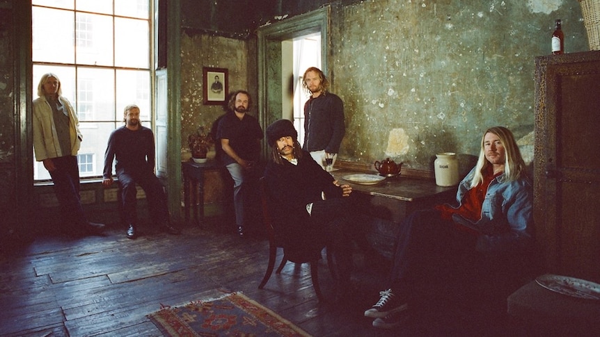 Six members of Ocean Alley sitting in a dark room with a dark wooden floor and decrepit green walls.