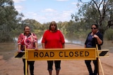 Three indigenous women stand in front of a road closed sign and behind them is a flooded park