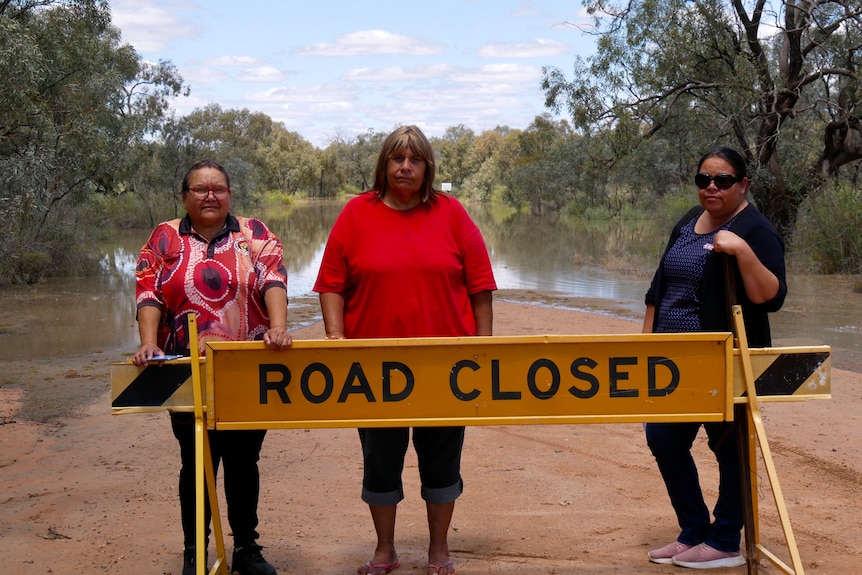 Three indigenous women stand in front of a road closed sign and behind them is a flooded park