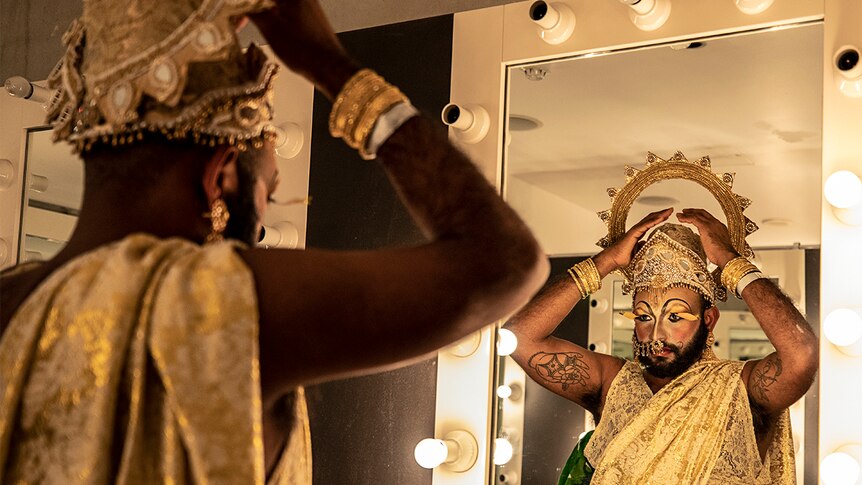 Colour photo of Shahmen Suku look at dressing room mirror and fixing costume head piece.
