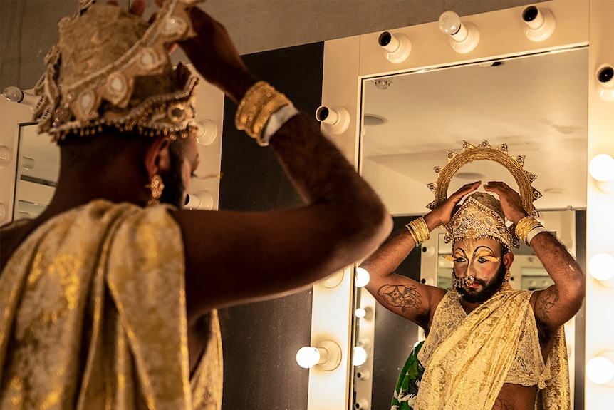 Colour photo of Shahmen Suku look at dressing room mirror and fixing costume head piece.