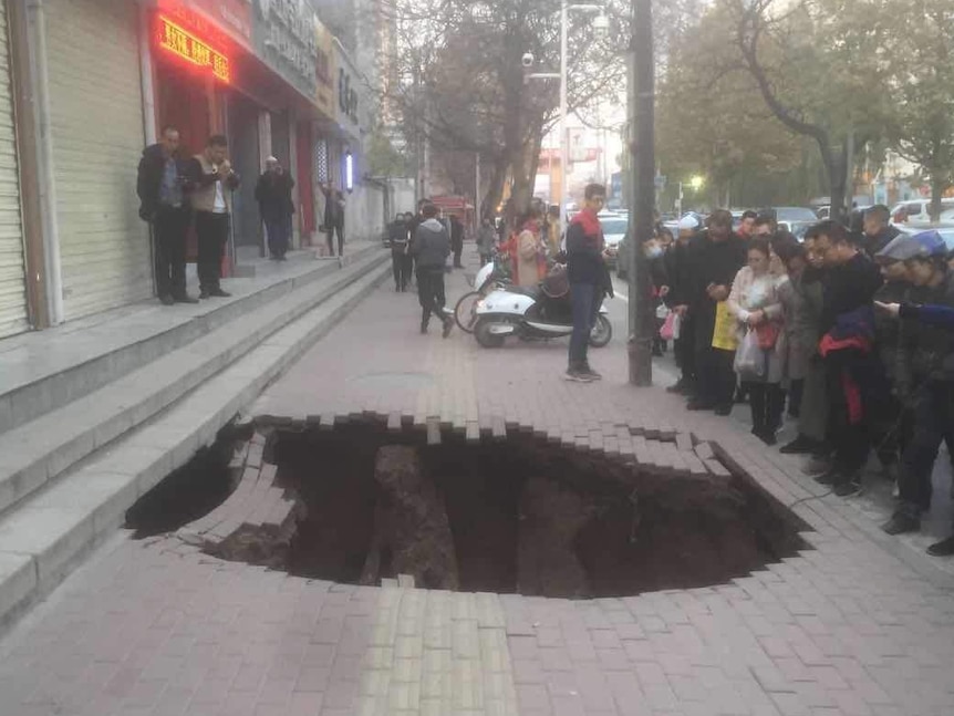 A crowd gathers around a large hole on a paved footpath