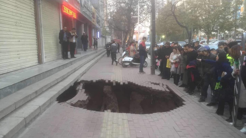 A crowd gathers around a large hole on a paved footpath