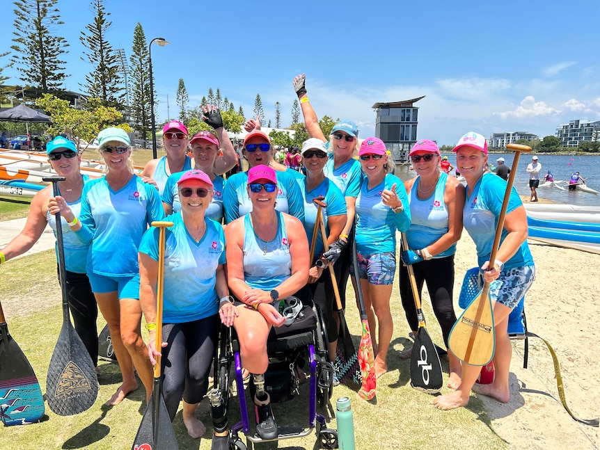 A group of women in blue team shirts and pink hats hold wooden paddles and pose in front of canoes