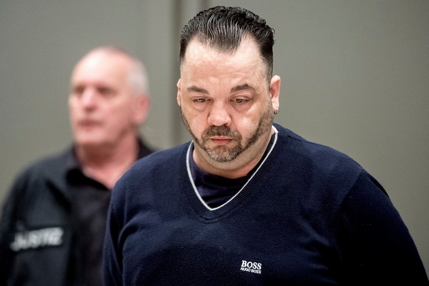 A man with a short beard and brushed-back hair looks at the ground as he stands in a court room.