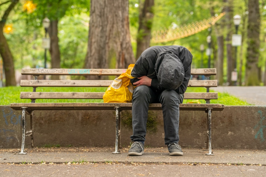Man crouched over on a park bench in Oregon with a yellow tote bag next to him.
