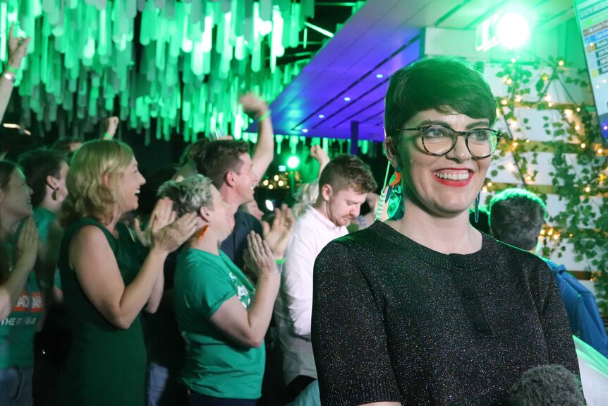 A woman smiles with green lights and people clapping behind her.