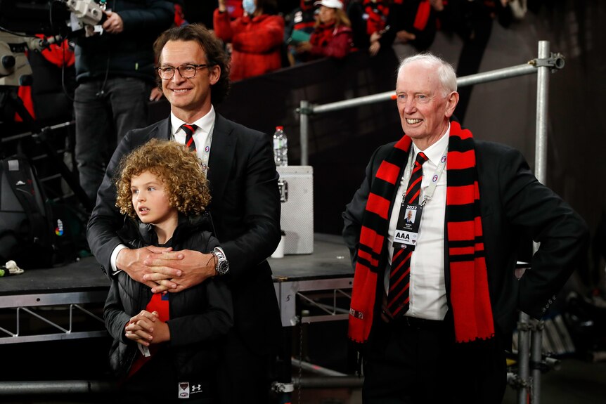 A man in a suit stands with his arms around a young girl, while next to him another man in a suit wearing an Essendon scarf.